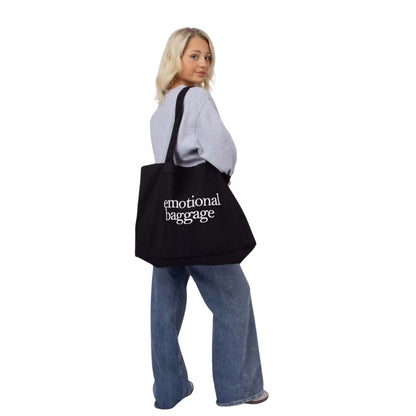 Person holding a black tote bag with 'emotional baggage' text on a white background