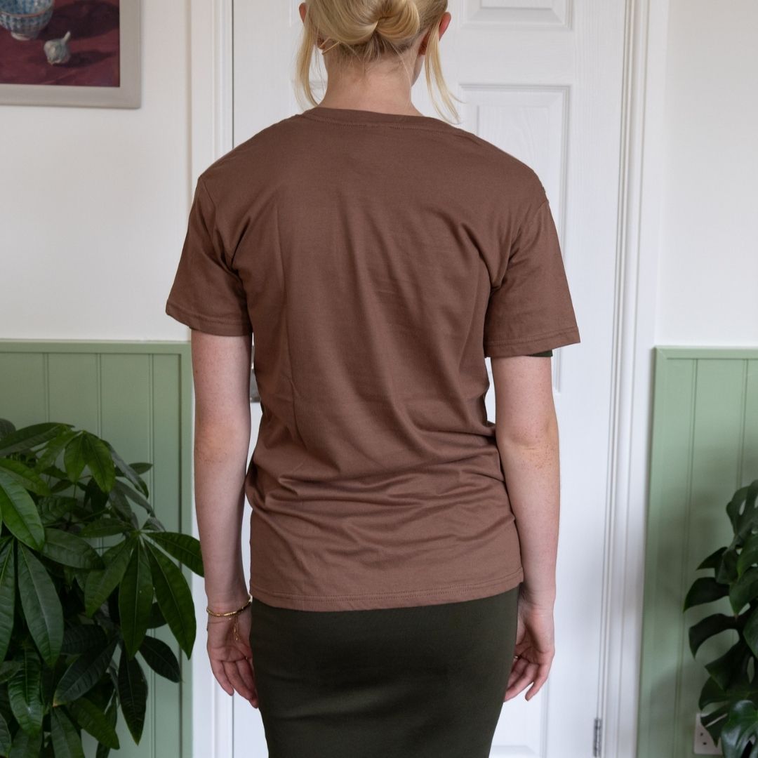 Person wearing a brown t-shirt standing indoors with plants in the background