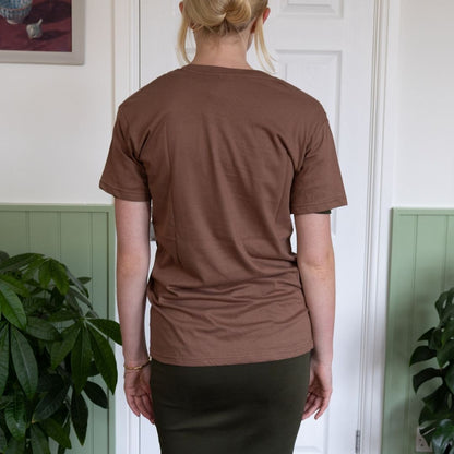 Person wearing a brown t-shirt standing indoors with plants in the background