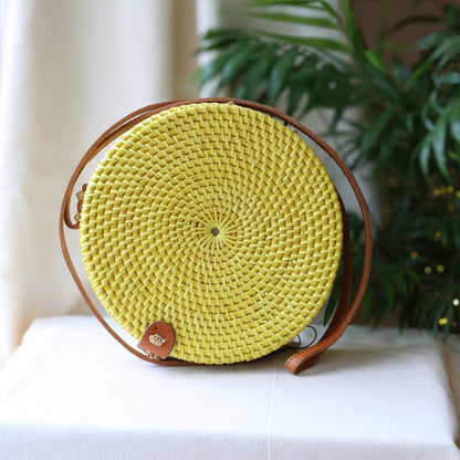 Round woven bag with a yellow center and brown leather strap on a white surface with a plant in the background.