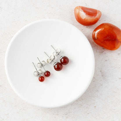 Two pairs of Carnelian stud earrings on a white trinket dish, with two red stones on a light surface.