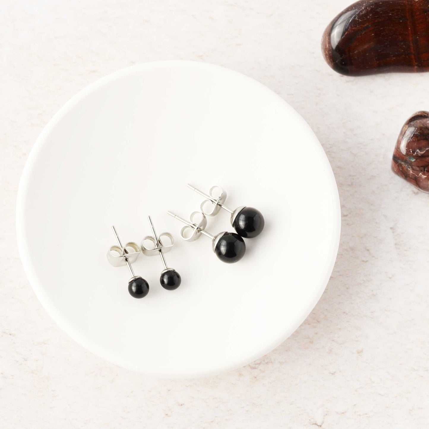 Black stud earrings on a white trinket dish with a dark stones in the background