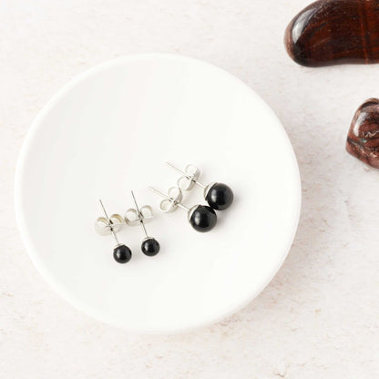 Black stud earrings on a white trinket dish with a dark stones in the background