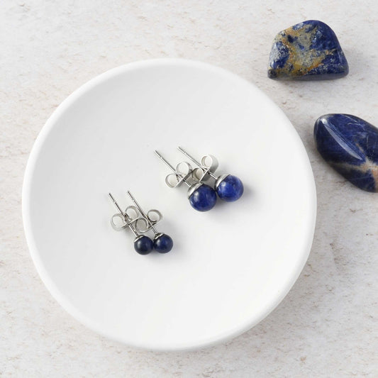 Two pairs of dark blue stone earrings on a white trinket dish with stones in the background