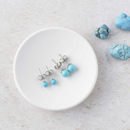 Two pairs of turquoise stud earrings on a white trinket dish with turquoise stones in the background.