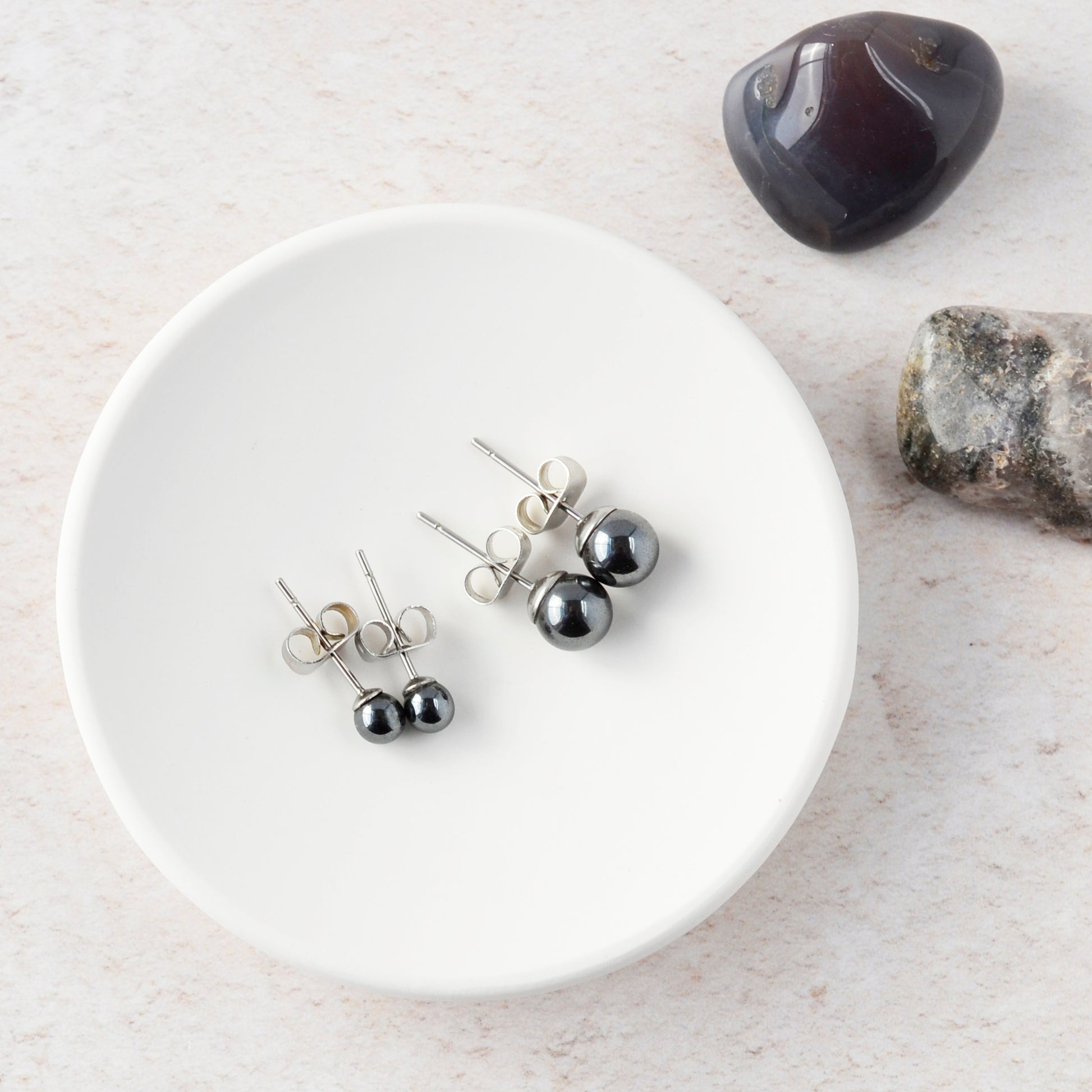 Two pairs of Hematite stud earrings on a white trinket dish with stones in the background