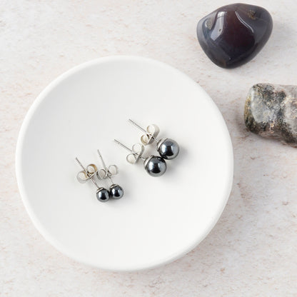 Two pairs of Hematite stud earrings on a white trinket dish with stones in the background