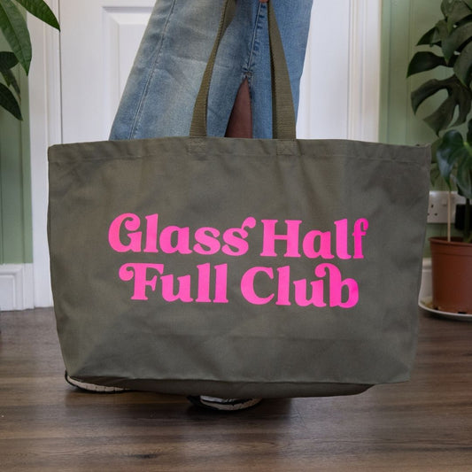 Person holding a green tote bag with pink text 'Glass Half Full Club' in an indoor setting.