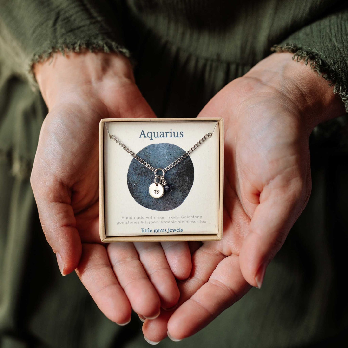 Hands holding a small square eco-friendly gift box containing a dainty Aquarius zodiac sign necklace