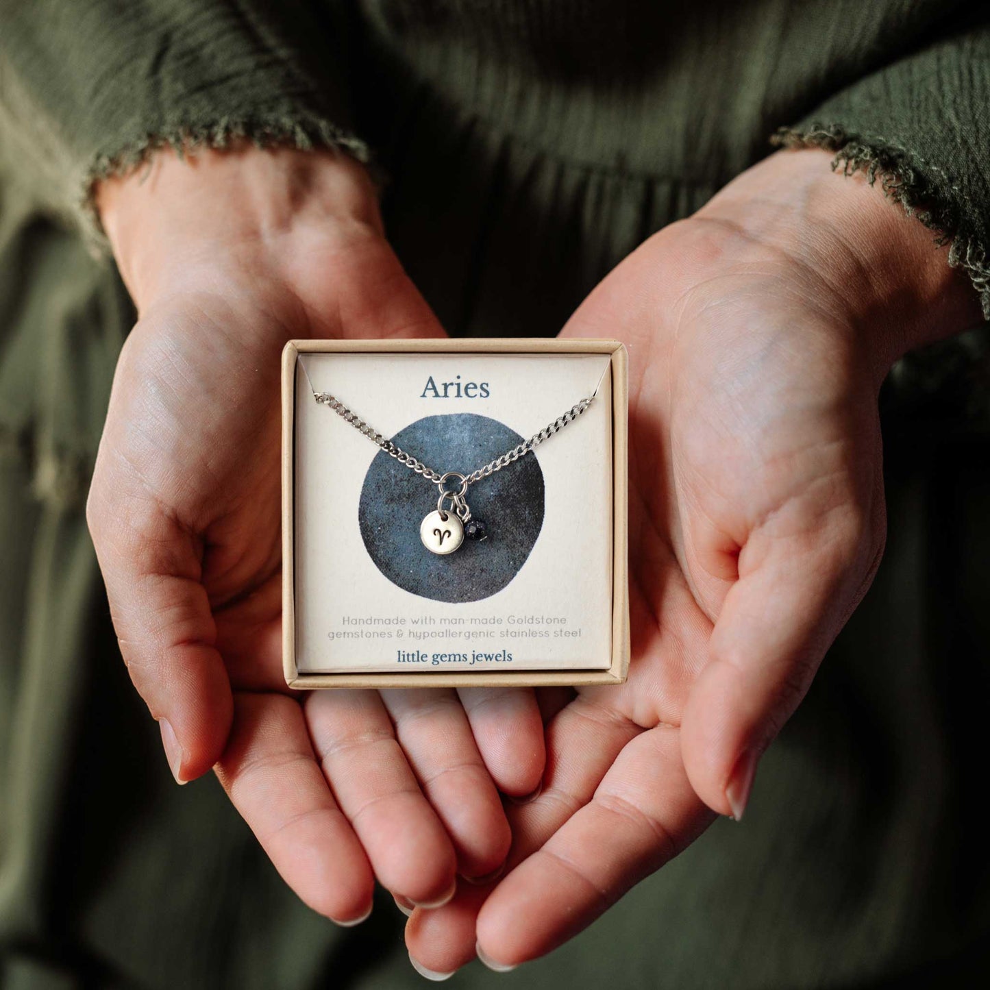 Hands holding a small square eco-friendly gift box containing a dainty Aries zodiac sign necklace