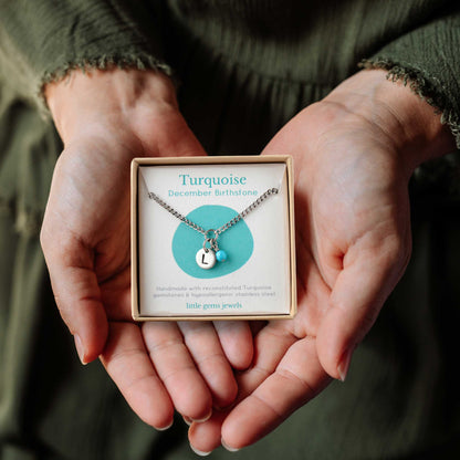 Hands holding a small square eco-friendly gift box containing a tiny initial and Turquoise December birthstone necklace