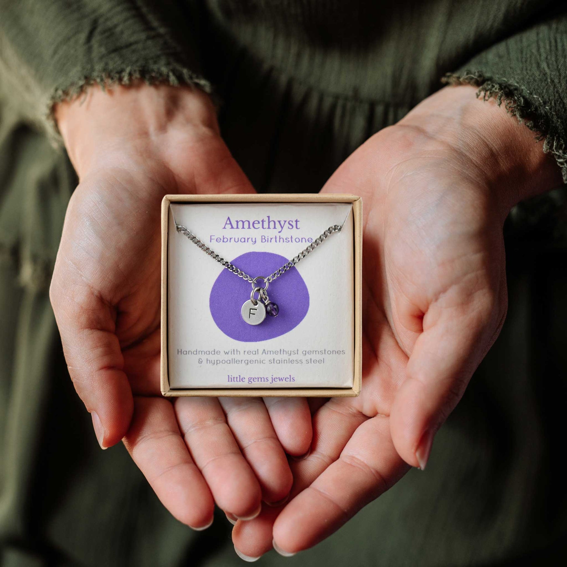 Hands holding a small square eco-friendly gift box containing a tiny initial and Amethyst February birthstone necklace