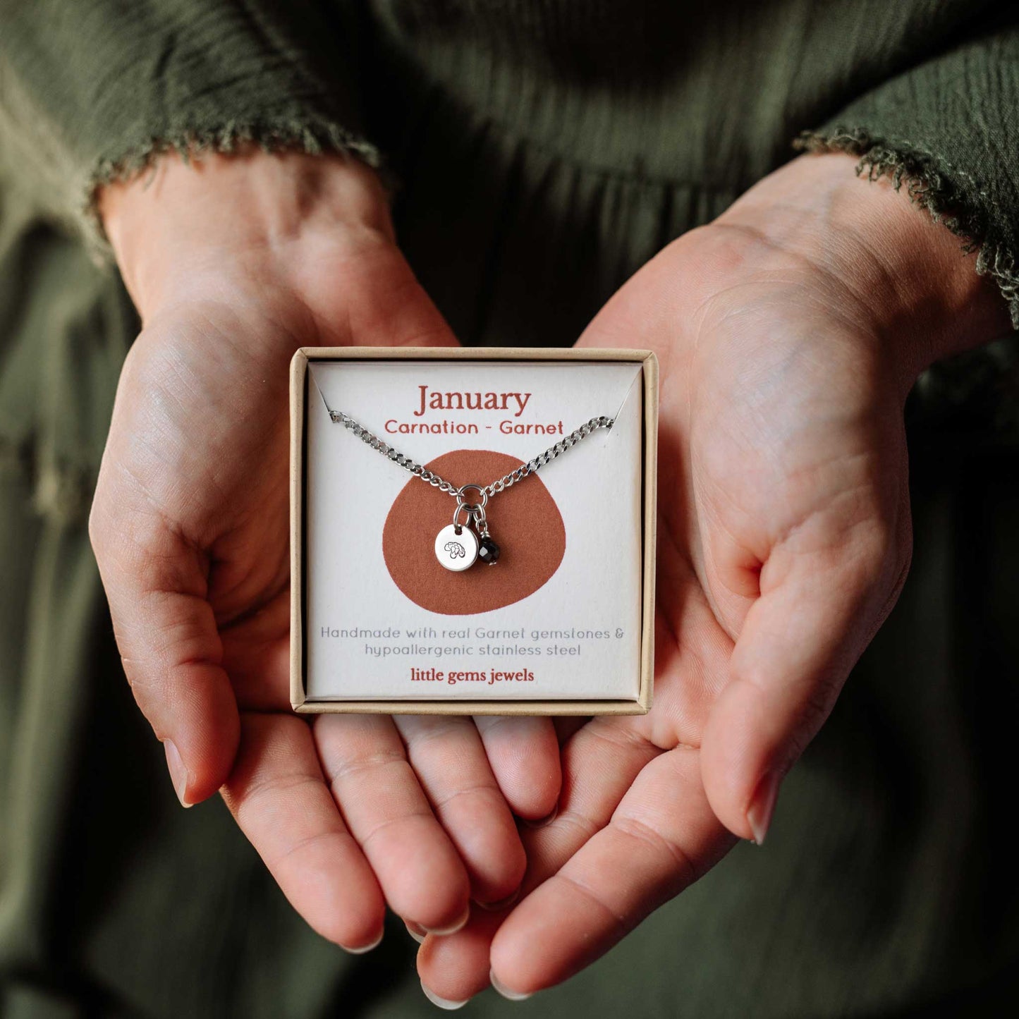 Woman's hands holding a small square eco-friendly gift box containing a dainty January birthflower with Garnet birthstone necklace