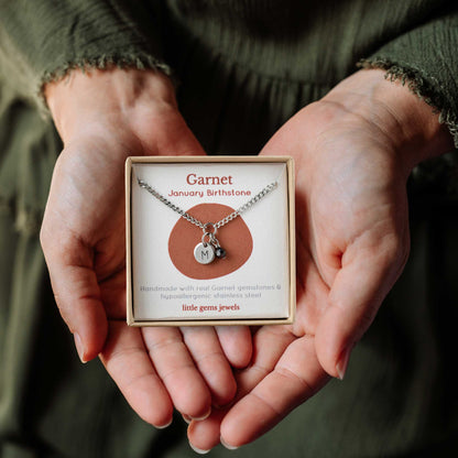 Hands holding a small square eco-friendly gift box containing a tiny initial and Garnet January birthstone necklace
