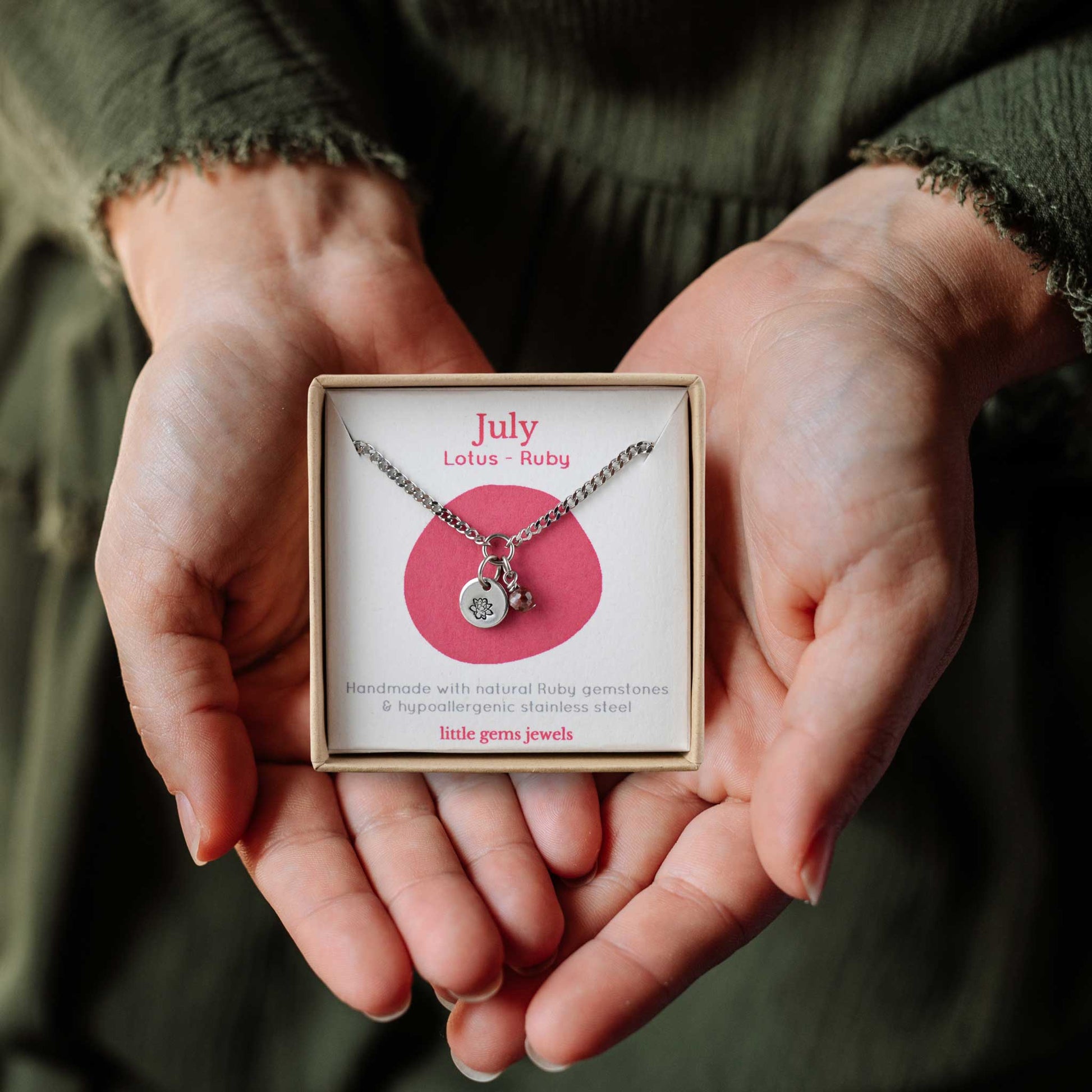 Woman's hands holding a small square eco-friendly gift box containing a dainty July birthflower with Ruby birthstone necklace