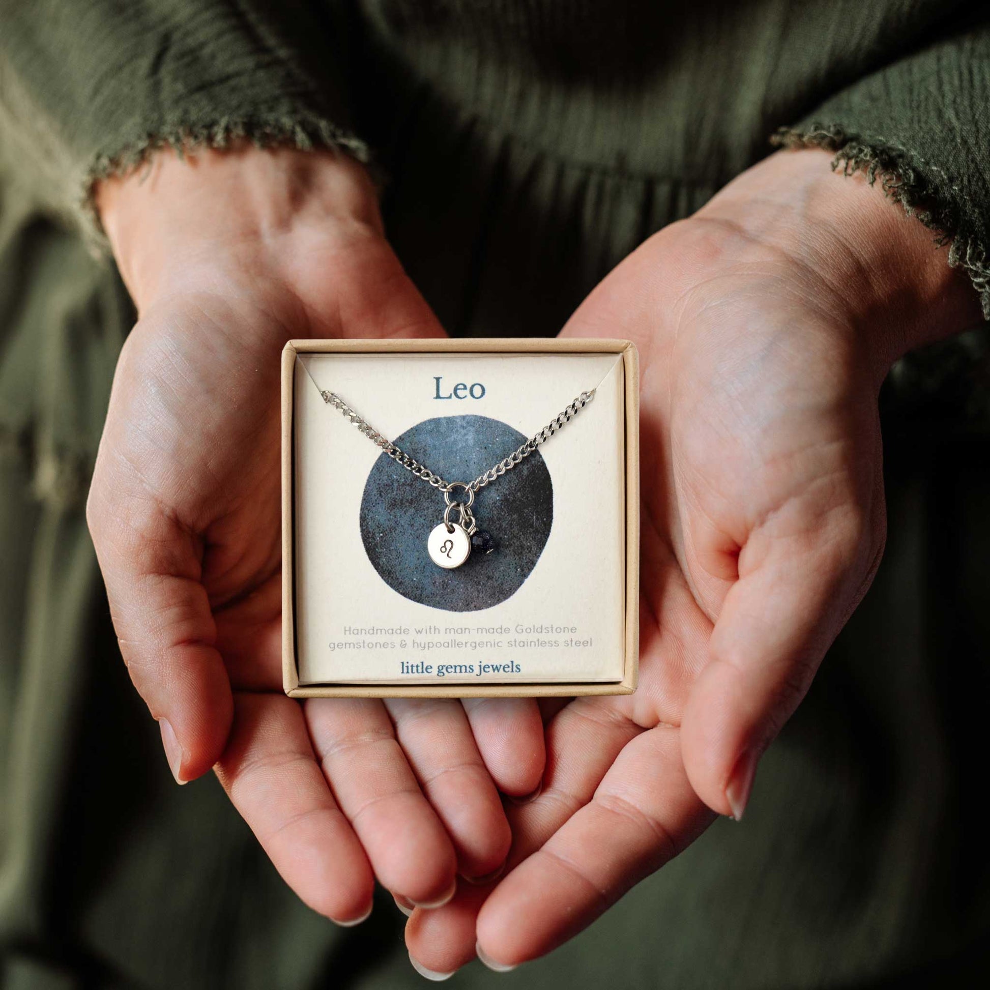 Hands holding a small square eco-friendly gift box containing a dainty Leo zodiac sign necklace