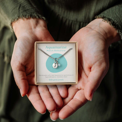 Hands holding a small square eco-friendly gift box containing a tiny initial and Aquamarine March birthstone necklace