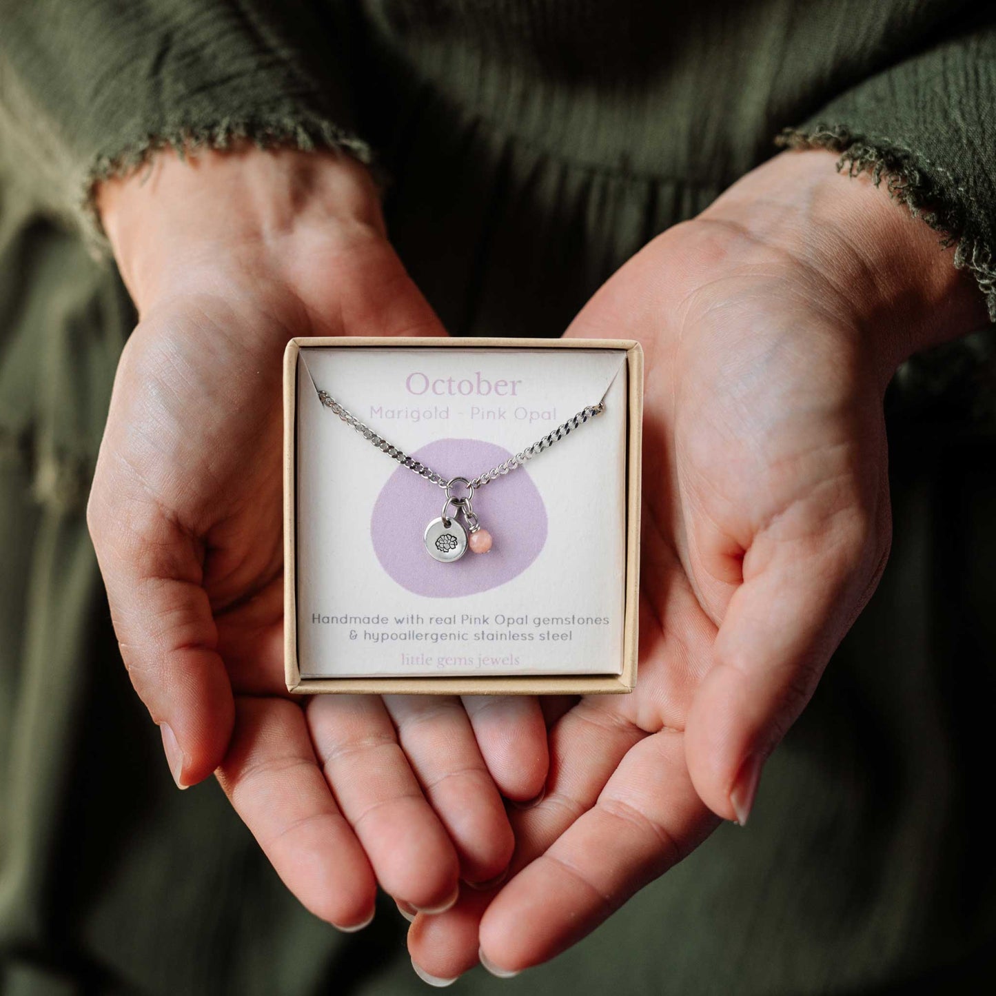 Woman's hands holding a small square eco-friendly gift box containing a dainty October birthflower with Pink Opal birthstone necklace