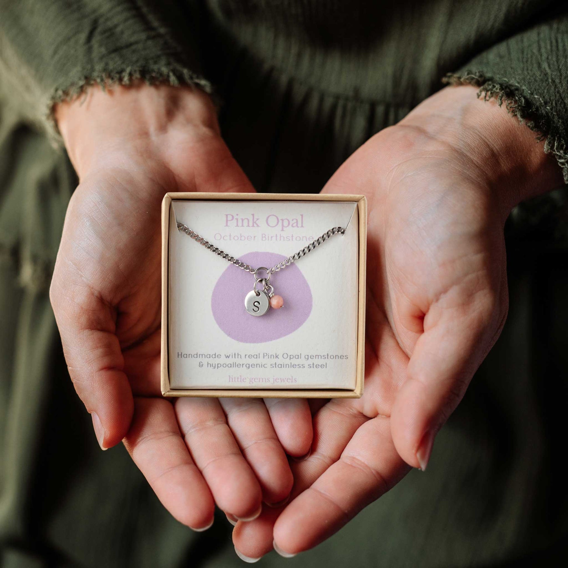 Hands holding a small square eco-friendly gift box containing a tiny initial and Pink Opal October birthstone necklace