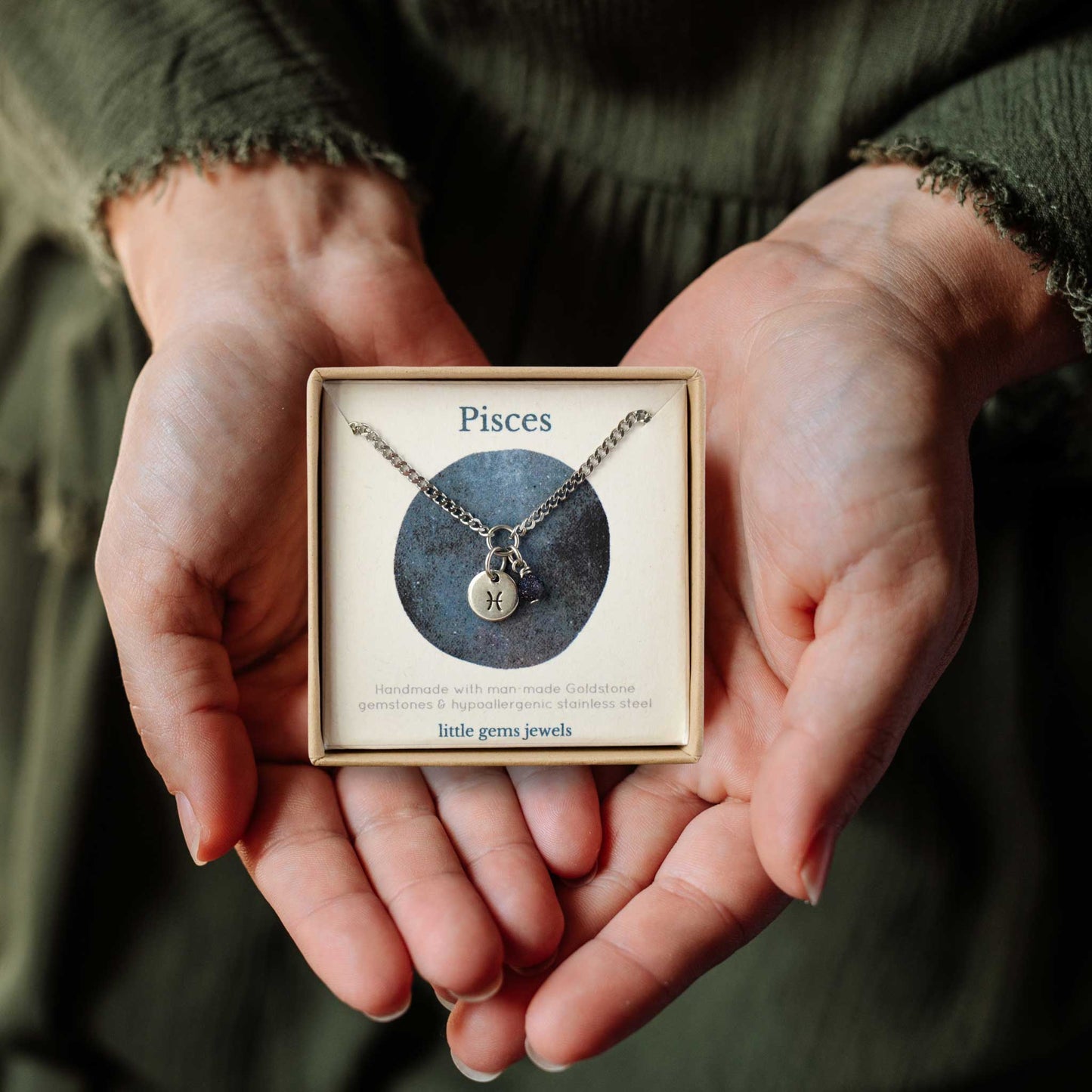 Hands holding a small square eco-friendly gift box containing a dainty Pisces zodiac sign necklace