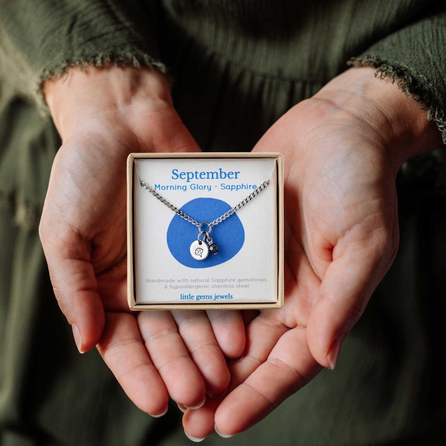 Woman's hands holding a small square eco-friendly gift box containing a dainty September birthflower with Sapphire birthstone necklace