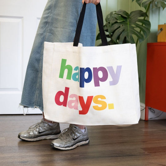Person holding a white tote bag with 'happy days' text in colorful letters.