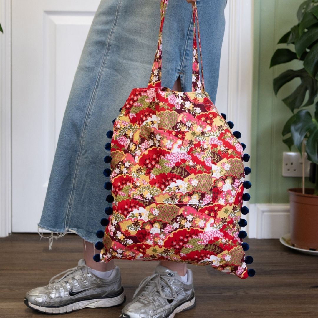 Person holding a red floral bag with pom-poms indoors