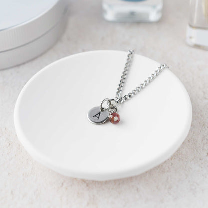 Tiny necklace with round disc pendant stamped with letter A and Ruby gemstone charm laying in a white trinket dish