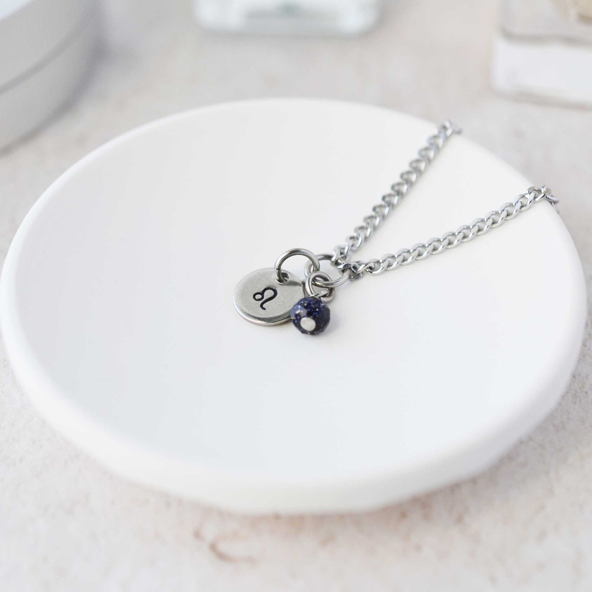 Tiny Leo zodiac sign disc pendant necklace with Blue Goldstone charm laying on white trinket dish