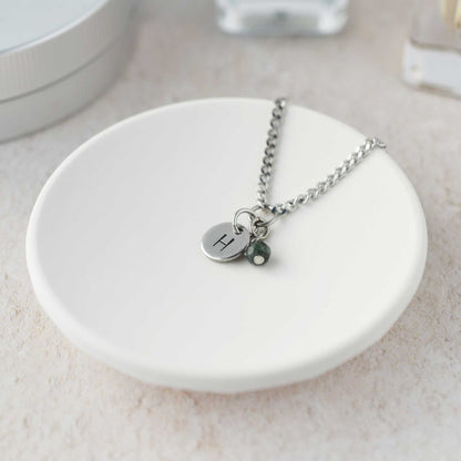 Tiny necklace with round disc pendant stamped with letter H and Emerald gemstone charm laying in a white trinket dish