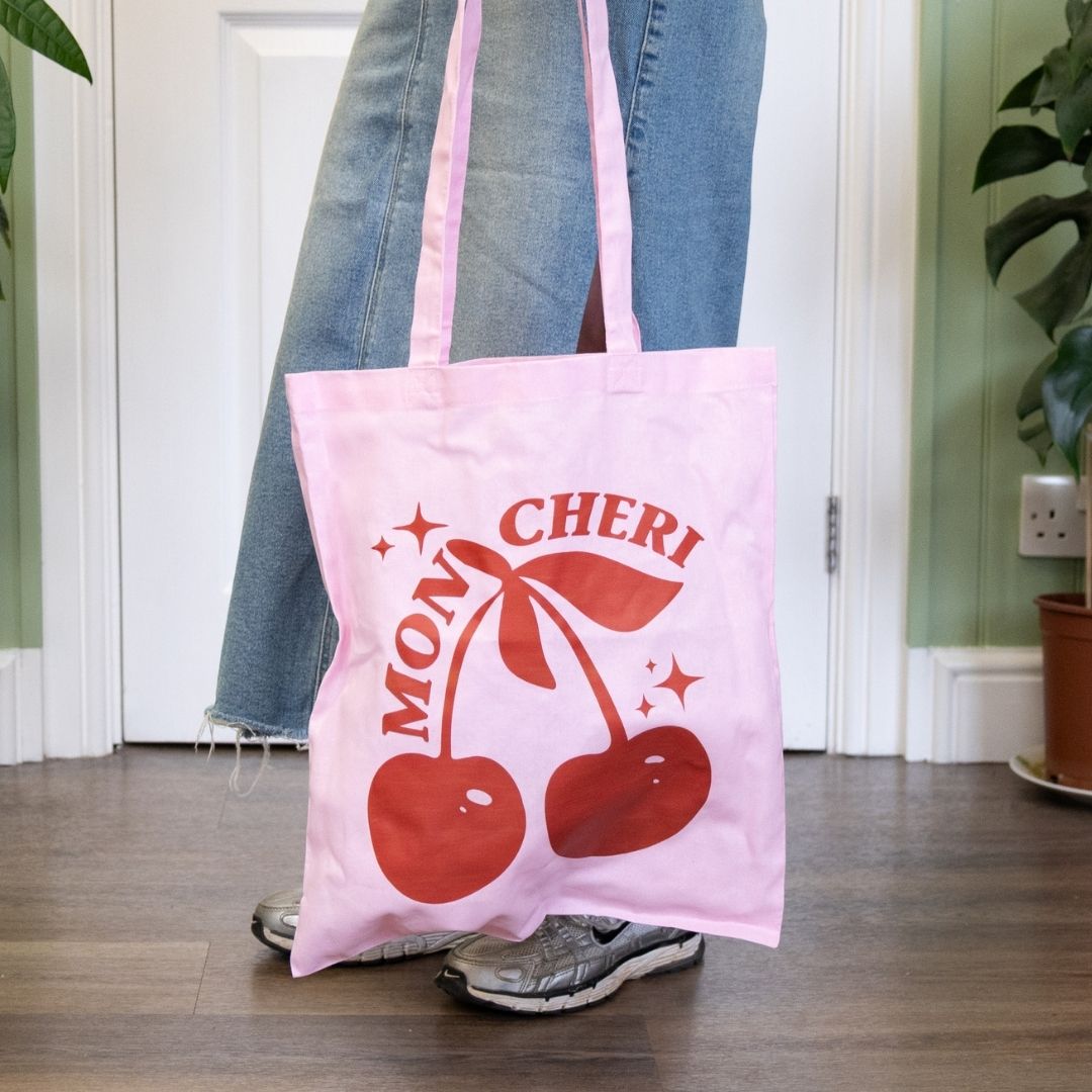 Person holding a pink tote bag with red cherries and 'Mon Cheri' text.