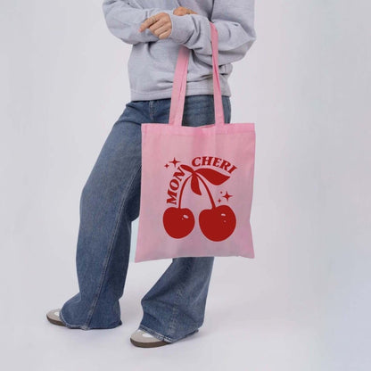 Person holding a pink tote bag with red cherries and 'Mon Cheri' text on a white background