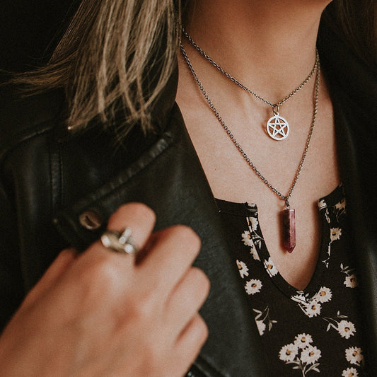 Person wearing a necklace with a pentacle pendant and another necklace with a purple pendant, against a dark background.