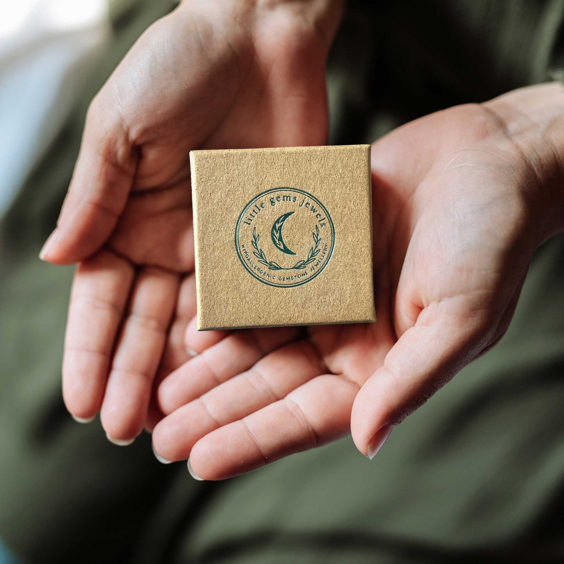 Hands holding a small square eco friendly gift box with Little Gems Jewels logo.