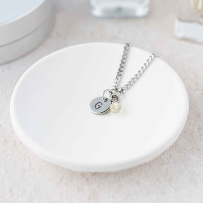 Tiny necklace with round disc pendant stamped with letter G and Citrine gemstone charm laying in a white trinket dish