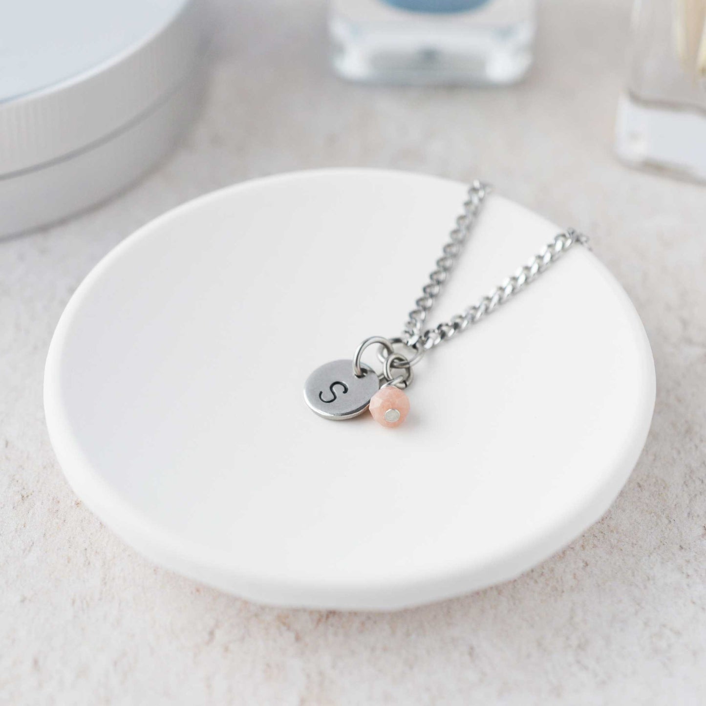 Tiny necklace with round disc pendant stamped with letter S and Pink Opal gemstone charm laying in a white trinket dish