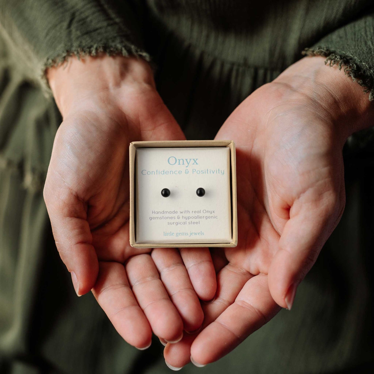 Hands holding small square gift box containing tiny Onyx stud earrings for confidence & positivity.