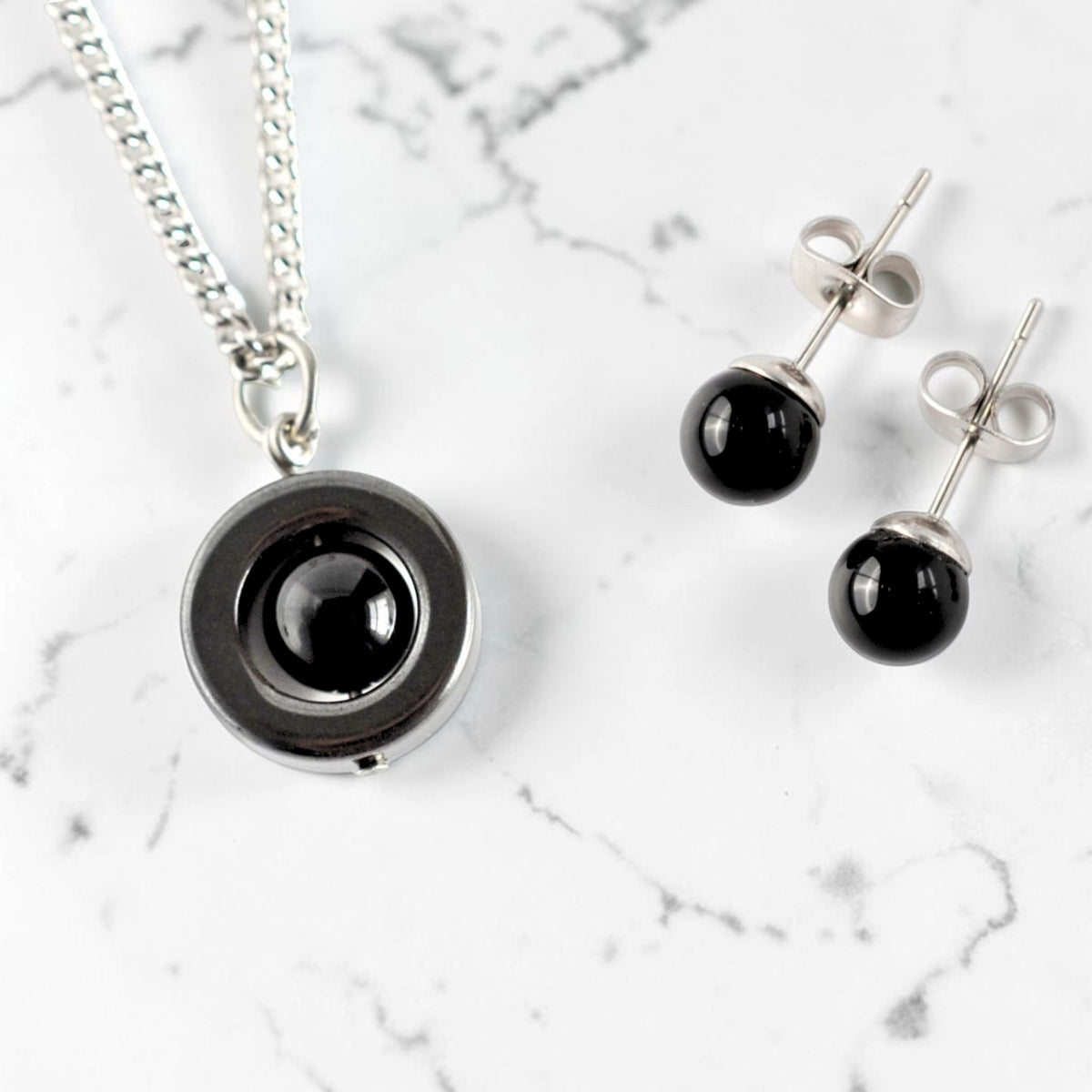 A steel necklace with a circular pendant featuring a glossy black Onyx gemstone, displayed alongside matching Onyx and surgical steel stud earrings on a white marble background.