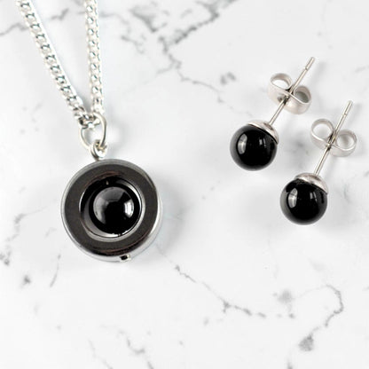 A steel necklace with a circular pendant featuring a glossy black Onyx gemstone, displayed alongside matching Onyx and surgical steel stud earrings on a white marble background.