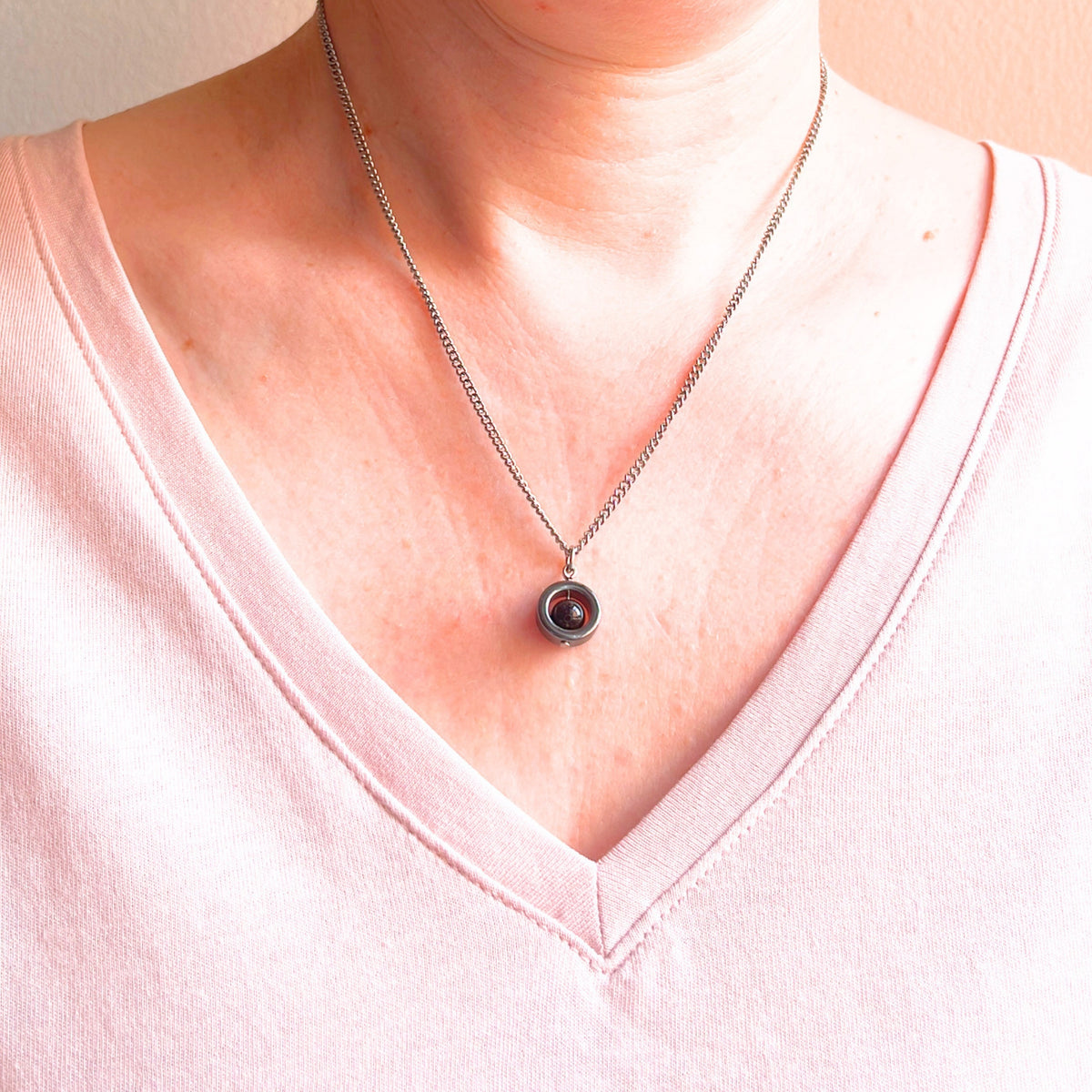 A close-up of a woman wearing a silver chain necklace with a circular pendant featuring a black gemstone center, styled with a light pink V-neck top.