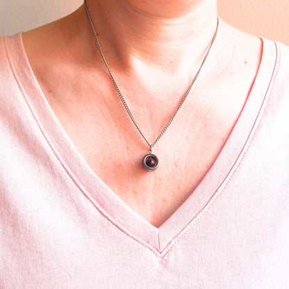 A close-up of a woman wearing a silver chain necklace with a circular pendant featuring a black gemstone center, styled with a light pink V-neck top.