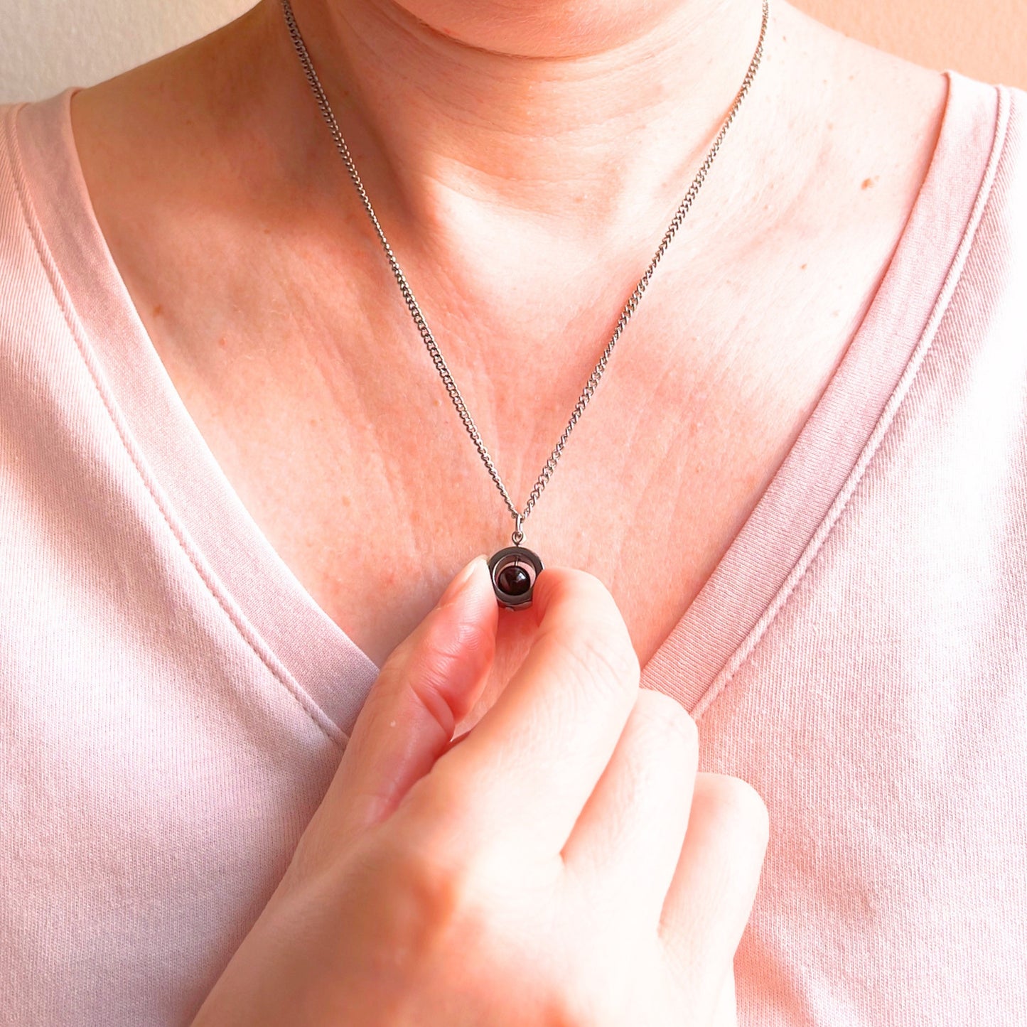 A close-up of a person wearing a silver chain necklace with a small dark gemstone pendant, with their hand touching the pendant. The person is wearing a light pink v-neck top.