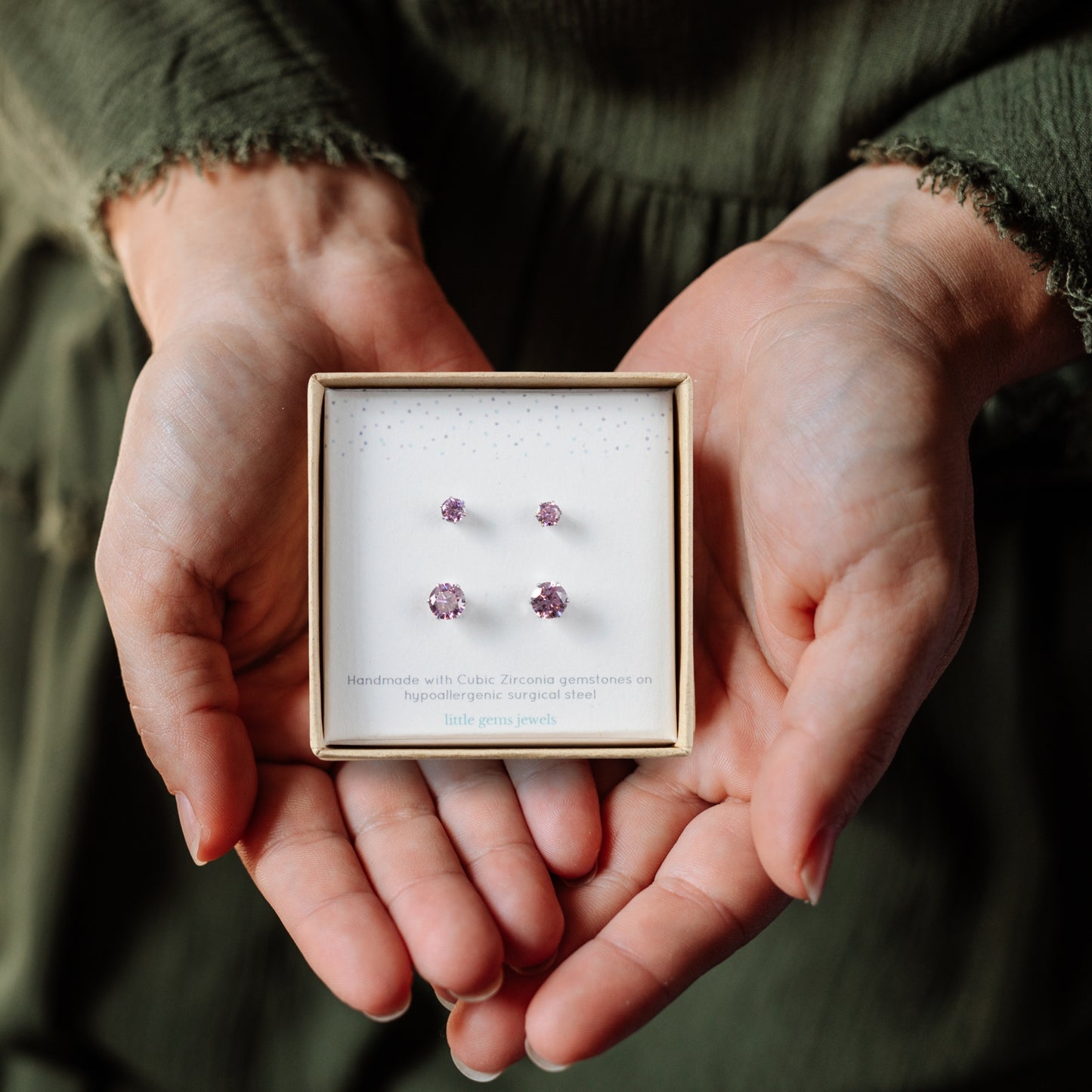 Hands holding a gift box with small and large pink CZ stud earrings inside.