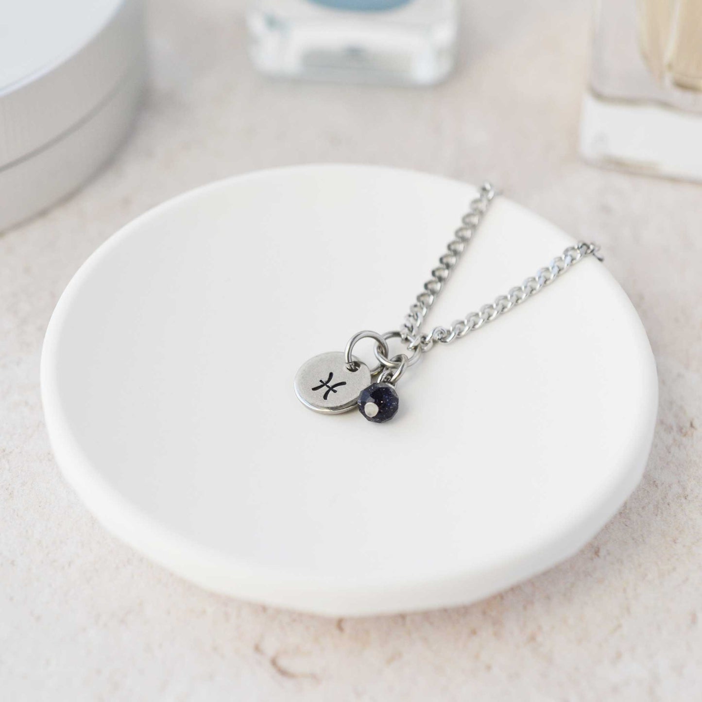 Tiny Pisces zodiac sign disc pendant necklace with Blue Goldstone charm laying on white trinket dish