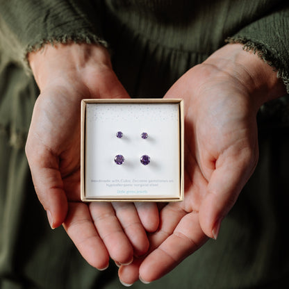 Hands holding a gift box with small and large purple CZ stud earrings inside.