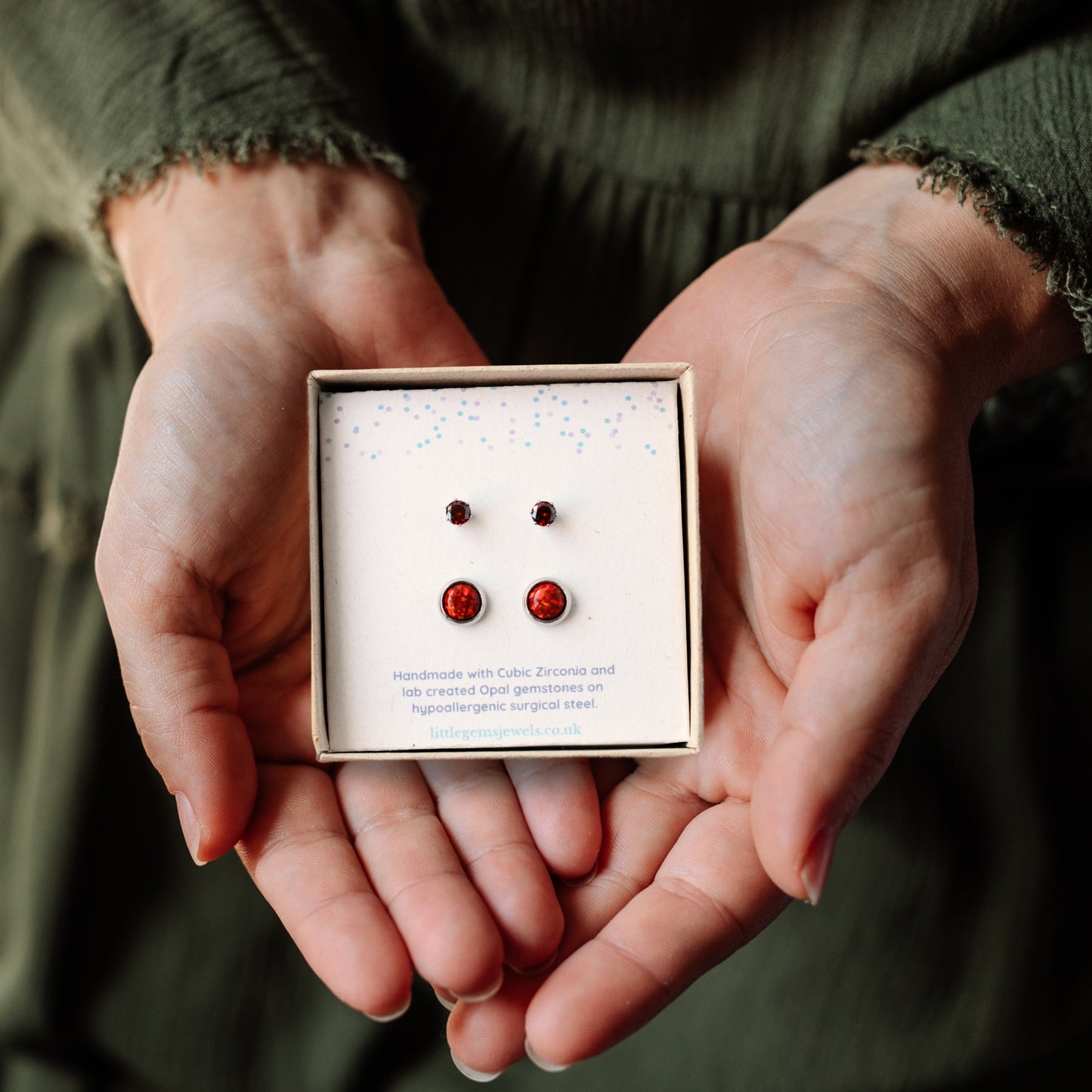 Hands holding a square gift box containing small and large red stone stud earrings.