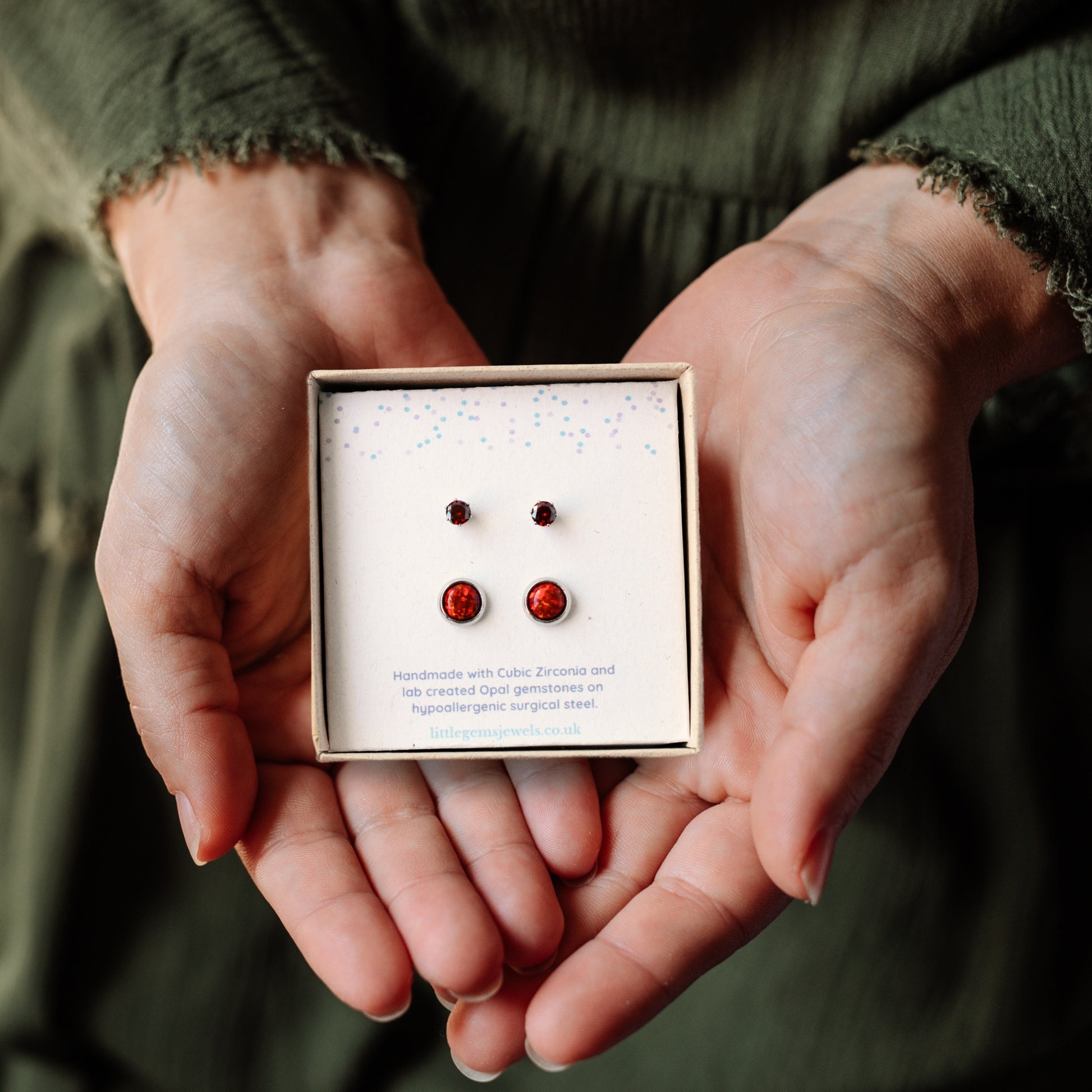 Hands holding a square gift box containing small and large red stone stud earrings.