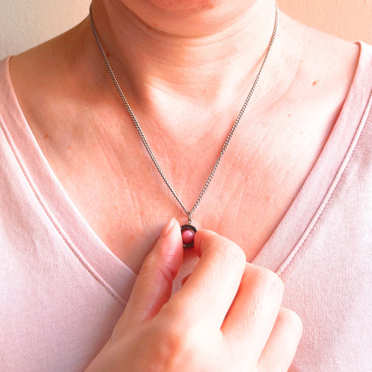 A close-up of a person wearing a silver chain necklace with a small dark pink gemstone pendant, with a hand spinning the pendant.