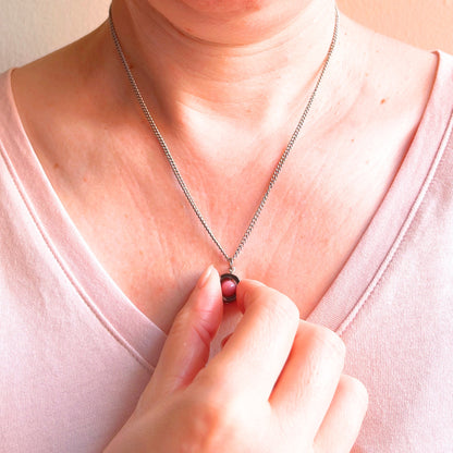 A close-up of a person wearing a silver chain necklace with a small dark pink gemstone pendant, with a hand spinning the pendant.