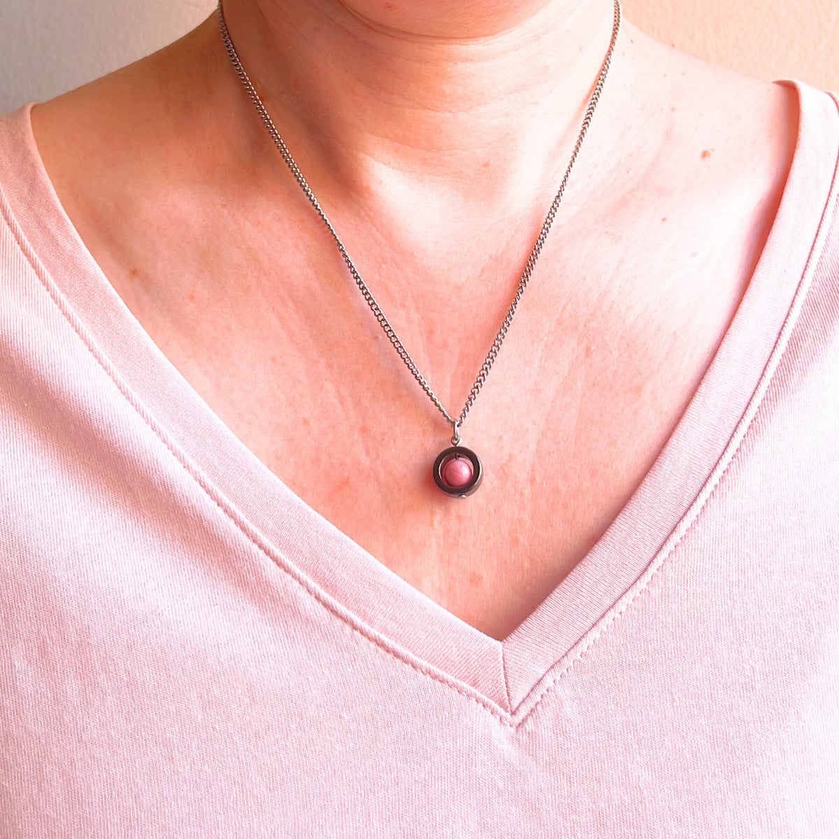 A close-up of a person's neck and chest wearing a delicate silver chain necklace with a circular pendant featuring a pink stone in the center, paired with a light pink V-neck top.